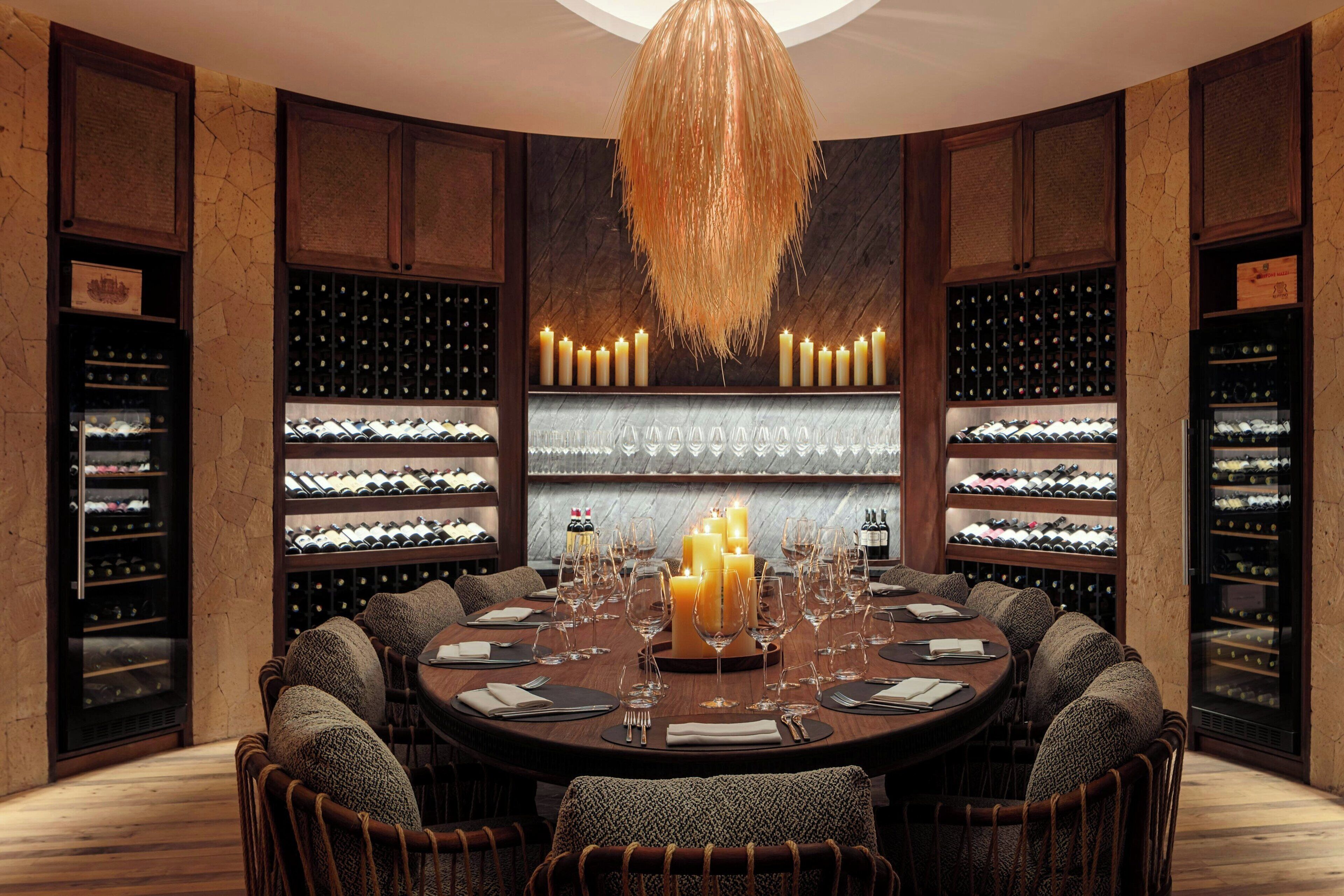 Una mesa redonda para comer con velas, botellas de vino y copas en una habitación con paredes y estantes de madera.