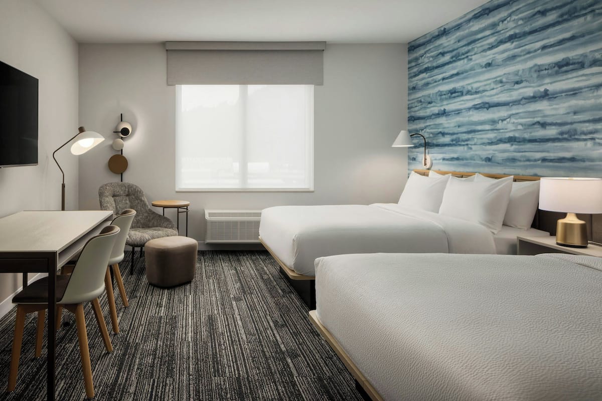 Habitación de hotel con cama, escritorio, silla, un pequeño otoman, televisor montado en la pared y un ventanal con vistas.