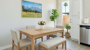 Un comedor con una mesa de madera, cuatro sillas, un florero y un cuadro de un paisaje montañoso.