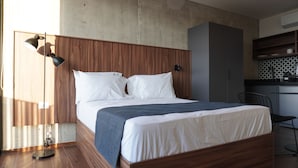 A modern bedroom with a wooden headboard, a bed with white linens, a blue throw blanket, a floor lamp, and a kitchen area with a black chair. - Lapso at Asís (Guadalajara)