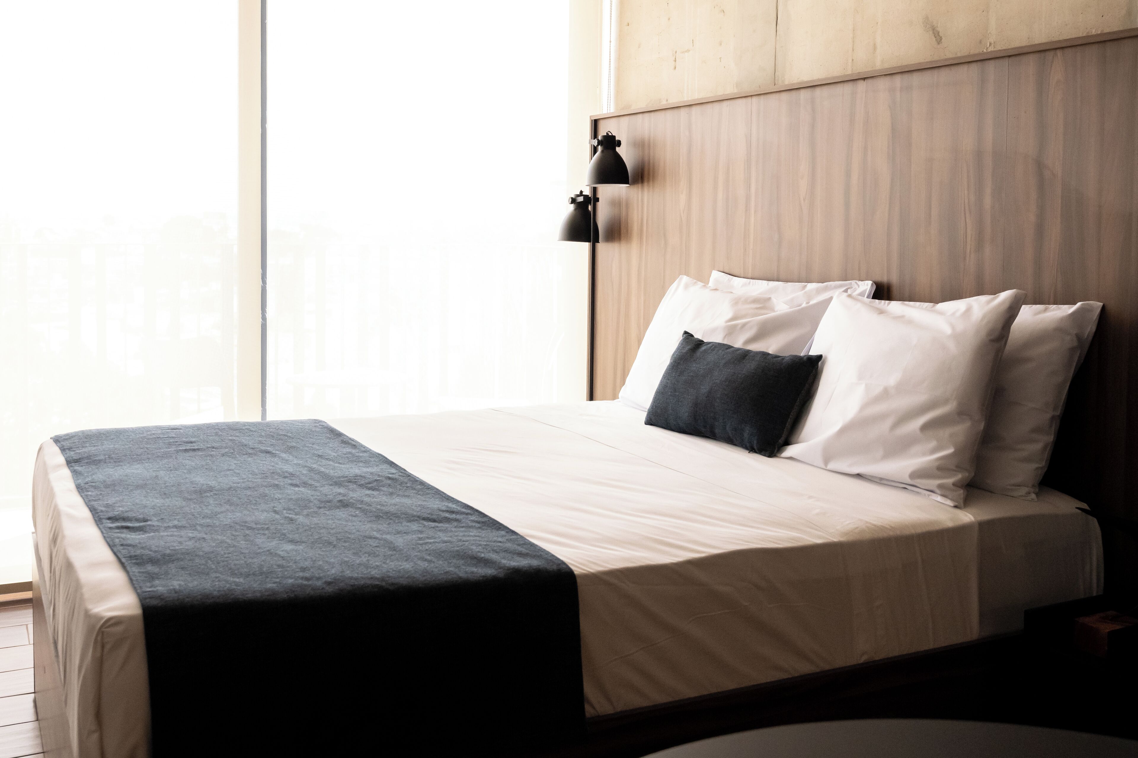 A neatly made bed with a dark throw, white pillows, and a wooden headboard in a room with large windows.