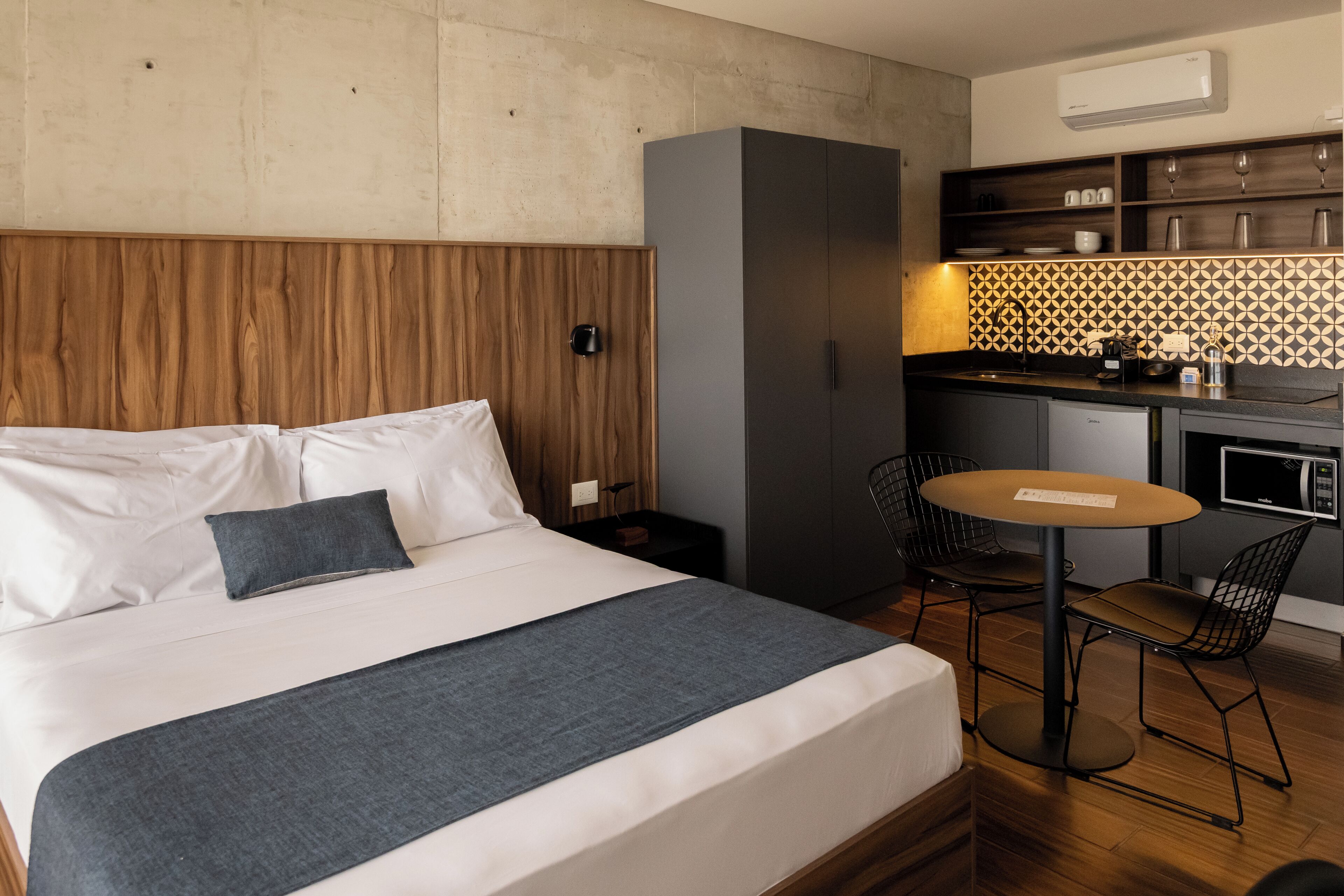 Habitación de hotel con cama, una mesita, dos sillas, kitchenette y cabecero de madera.