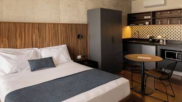 Habitación de hotel con cama, una mesita, dos sillas, kitchenette y cabecero de madera.