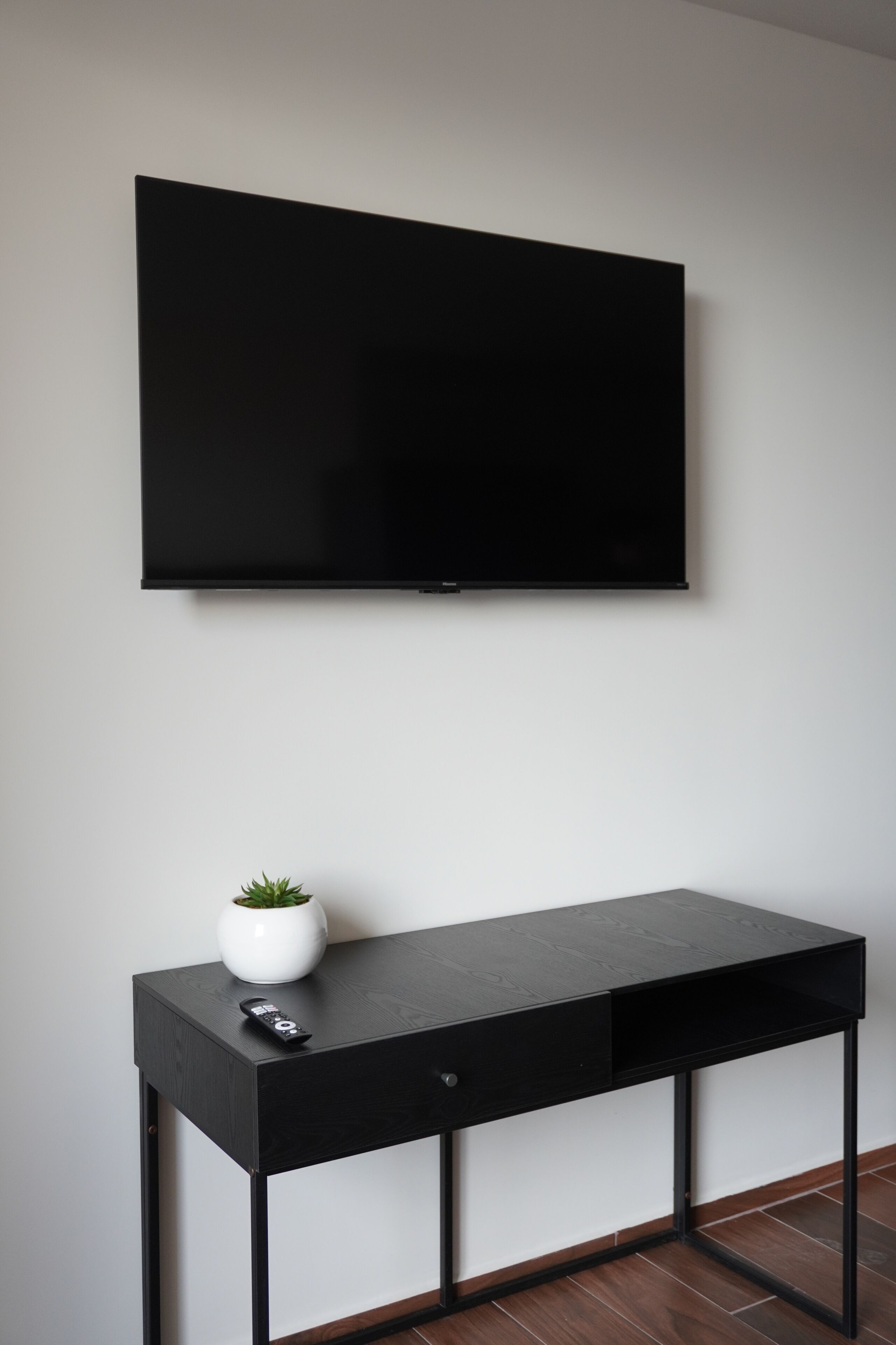 A modern room with a black TV mounted on the wall, a black desk with a remote control and a small plant on it, and a wooden floor.