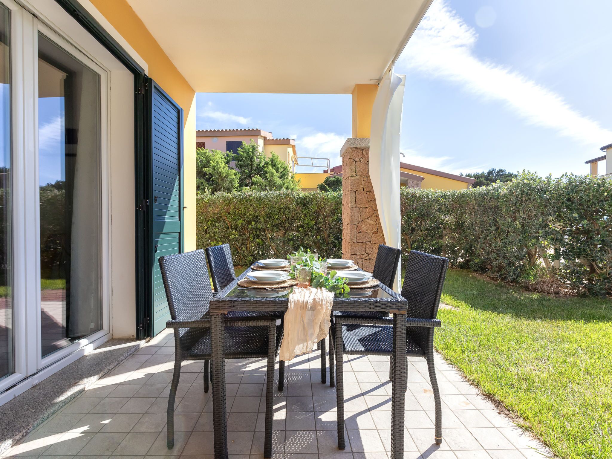 An outdoor patio with a table set for four, surrounded by wicker chairs, a stone pillar, and a white curtain.
