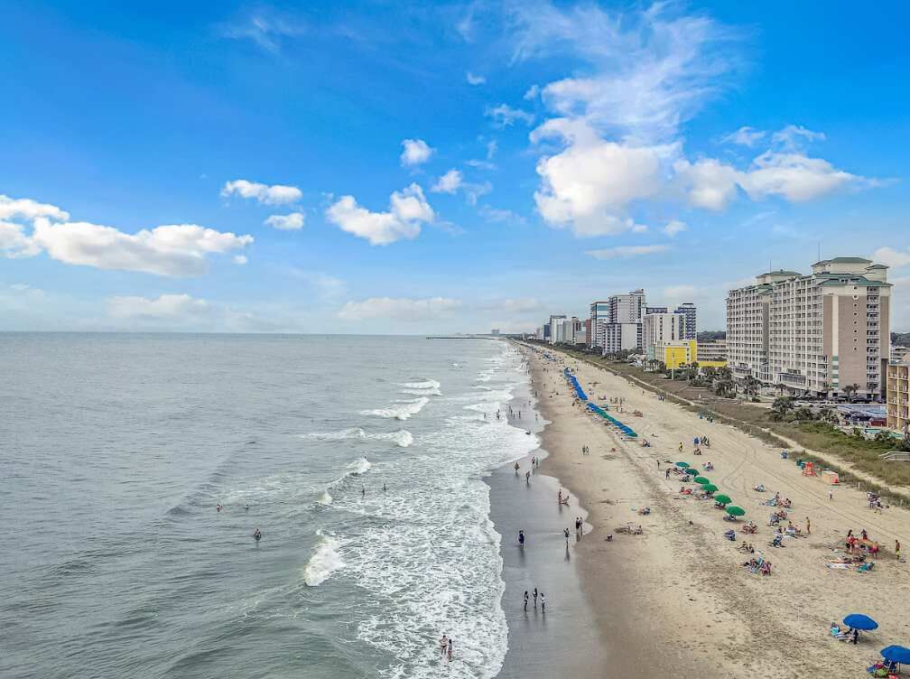 Un hôtel situé en bord de mer, avec une longue plage de sable, une vue sur l’océan et plusieurs immeubles de grande hauteur en arrière-plan.