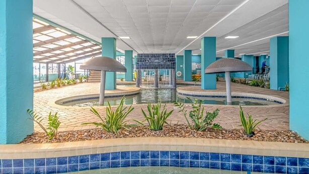 An indoor pool area with a central water feature, surrounded by tiled flooring and greenery.