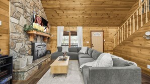 A cozy cabin interior with a stone fireplace, a gray sectional sofa, and a wooden ceiling. - 5BR, Heated Pool/Spa + Mtn View, Games, Karaoke, Firepit, 15 Min to PF (Sevierville)