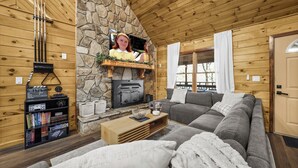 A cozy log cabin interior with a stone fireplace, a bookshelf, a gray sofa, and a wooden coffee table. - 5BR, Heated Pool/Spa + Mtn View, Games, Karaoke, Firepit, 15 Min to PF (Sevierville)