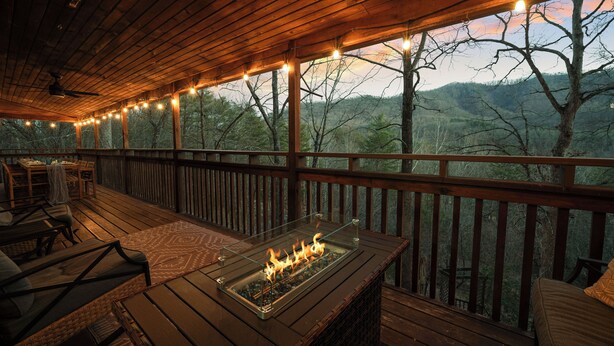 A wooden deck with a fire feature, outdoor seating, and a view of the surrounding forest. - 5BR, Heated Pool/Spa + Mtn View, Games, Karaoke, Firepit, 15 Min to PF (Sevierville)