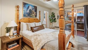 A four-poster bed with a wooden headboard, a nightstand with a lamp, a potted plant, and a painting on the wall.