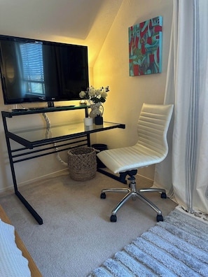 Un espace de bureau moderne avec un bureau en verre, une chaise de bureau blanche, un téléviseur, un vase décoratif avec des fleurs et une peinture abstraite colorée accrochée au mur.