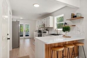 A modern kitchen with white cabinets, a large island with stools, and a view of the outdoors through a glass door.