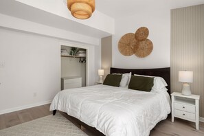 Quarto com uma cama grande, roupa de cama branca, mesa de cabeceira com abajur e um objeto decorativo na parede.