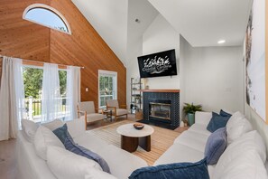 A spacious living room with a fireplace, a television mounted on the wall, and a large window overlooking a deck. - Club Paradise: Pet Friendly, Soundside, Newly Updated! (Nags Head)