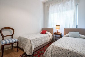 A bedroom with two single beds, a wooden chair, a nightstand with a lamp, and a window with sheer curtains.