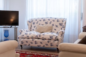 A living room with a floral-patterned armchair, a flat-screen TV, and a blue vase on a side table.