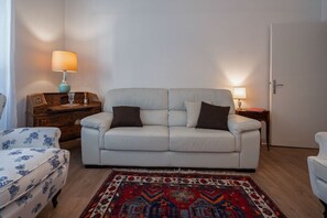 Ein Wohnzimmer mit einem weißen Sofa, zwei Beistelltischen mit Lampen, einem gemusterten Teppich und einer Holzkommode.