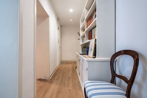 Un pasillo estrecho con piso de madera, un armario blanco lleno de libros y una silla de madera con cojín a rayas.