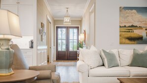 Un salón bien iluminado con un sofá blanco, una mesa redonda y una lámpara. Una puerta de madera da a otra habitación, y hay un cuadro en la pared.