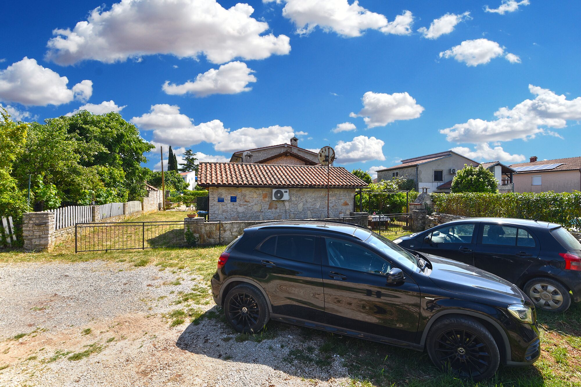 Un SUV noir était garé sur une allée de gravier, dans une zone rurale où se trouvaient des maisons en pierre et où le ciel était d’un bleu éclatant.