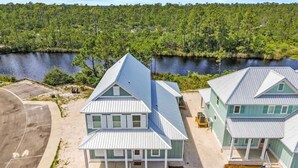 A two-story building with a gabled roof, located near a body of water with a forested background. - Tranquil Tides Orange Beach Getaway (Orange Beach)