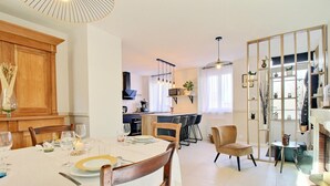 Un coin repas avec une table ronde dressée pour quatre personnes, une cuisine en arrière-plan et un espace de vie comprenant un canapé et une table d’appoint.