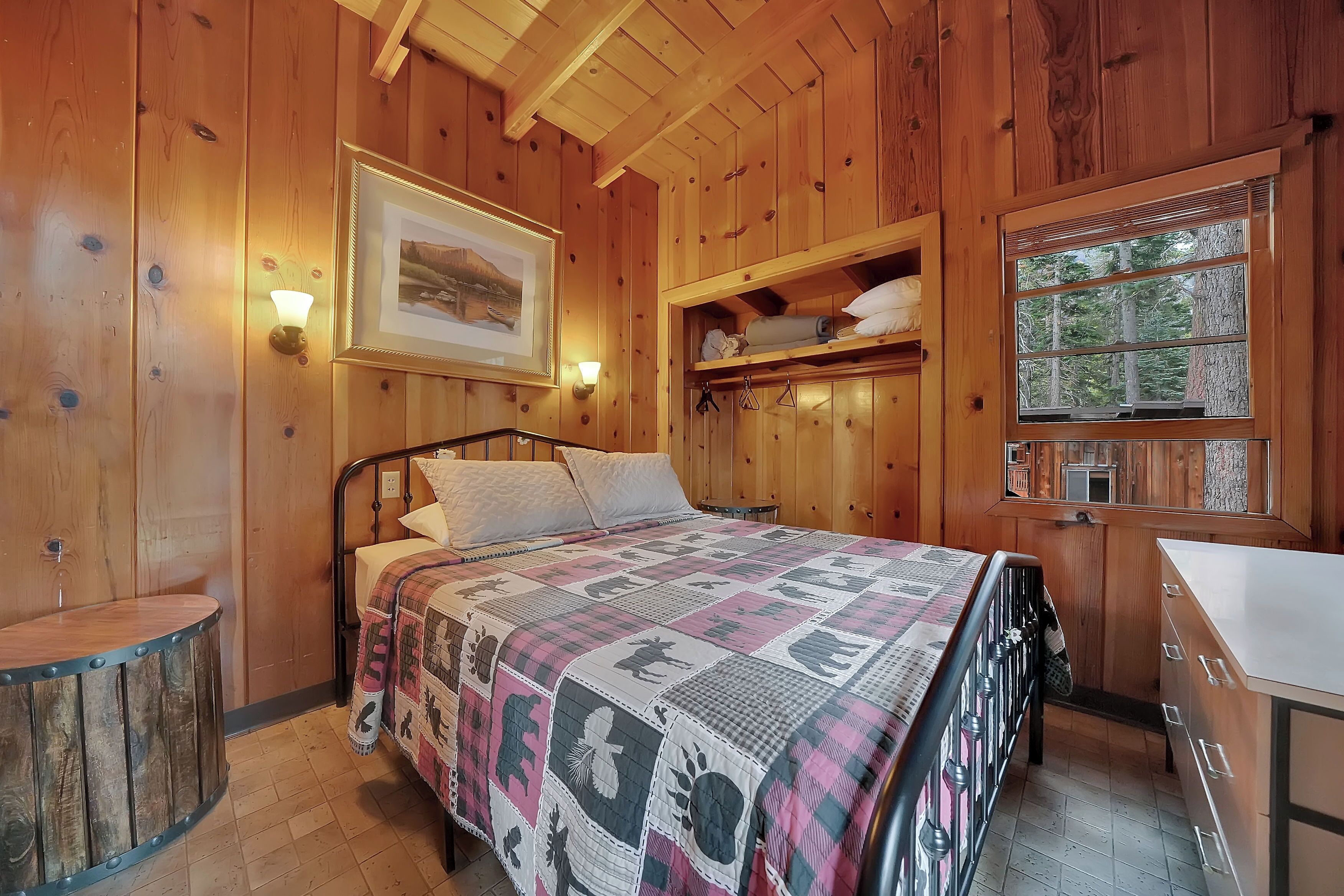 Une chambre dans une cabane en bois, avec un lit, une table de chevet, une étagère avec des serviettes, un tableau encadré au mur et une fenêtre donnant sur les arbres.