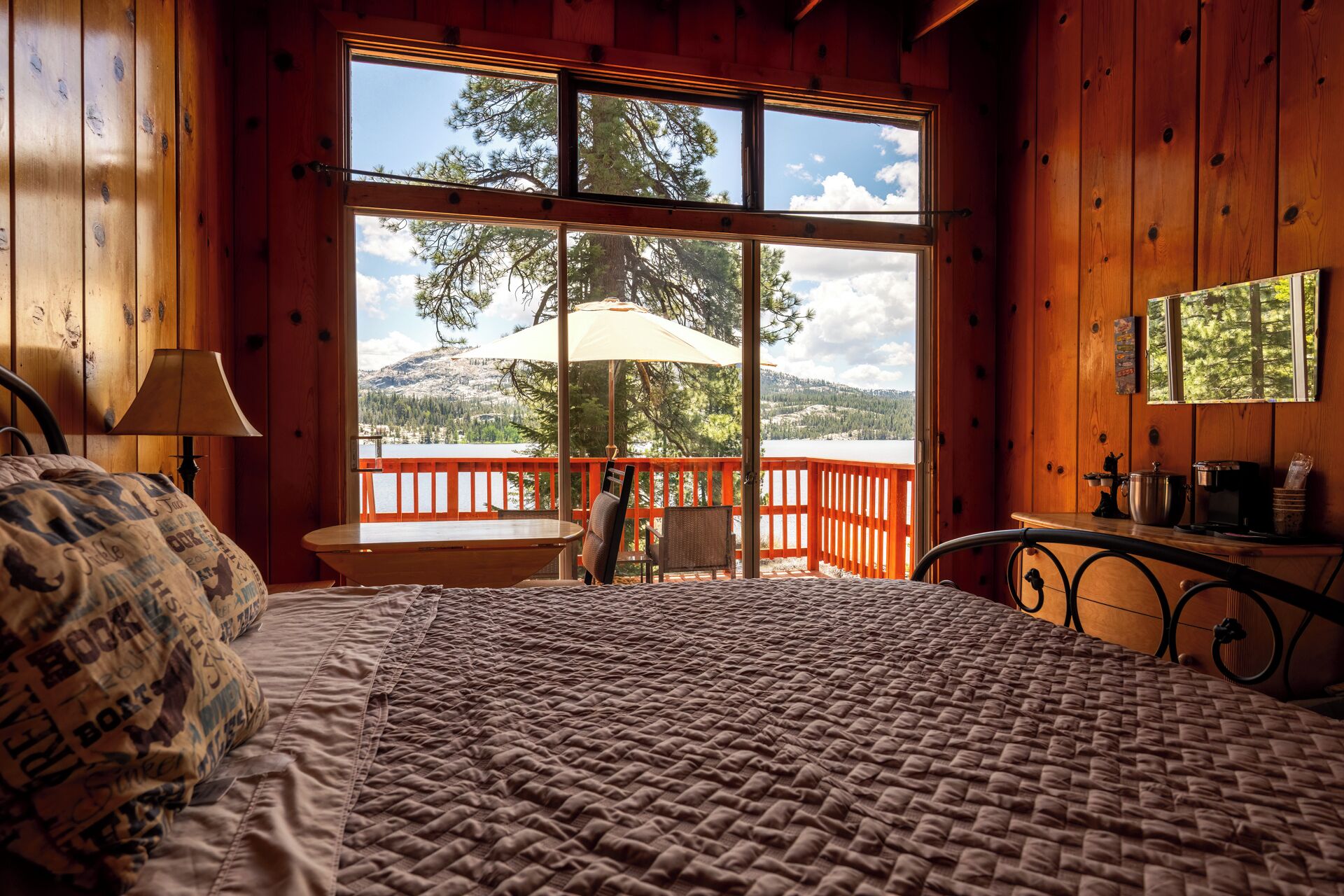 A bedroom with a large window, a bed, a nightstand, and a view of a lake and mountains.