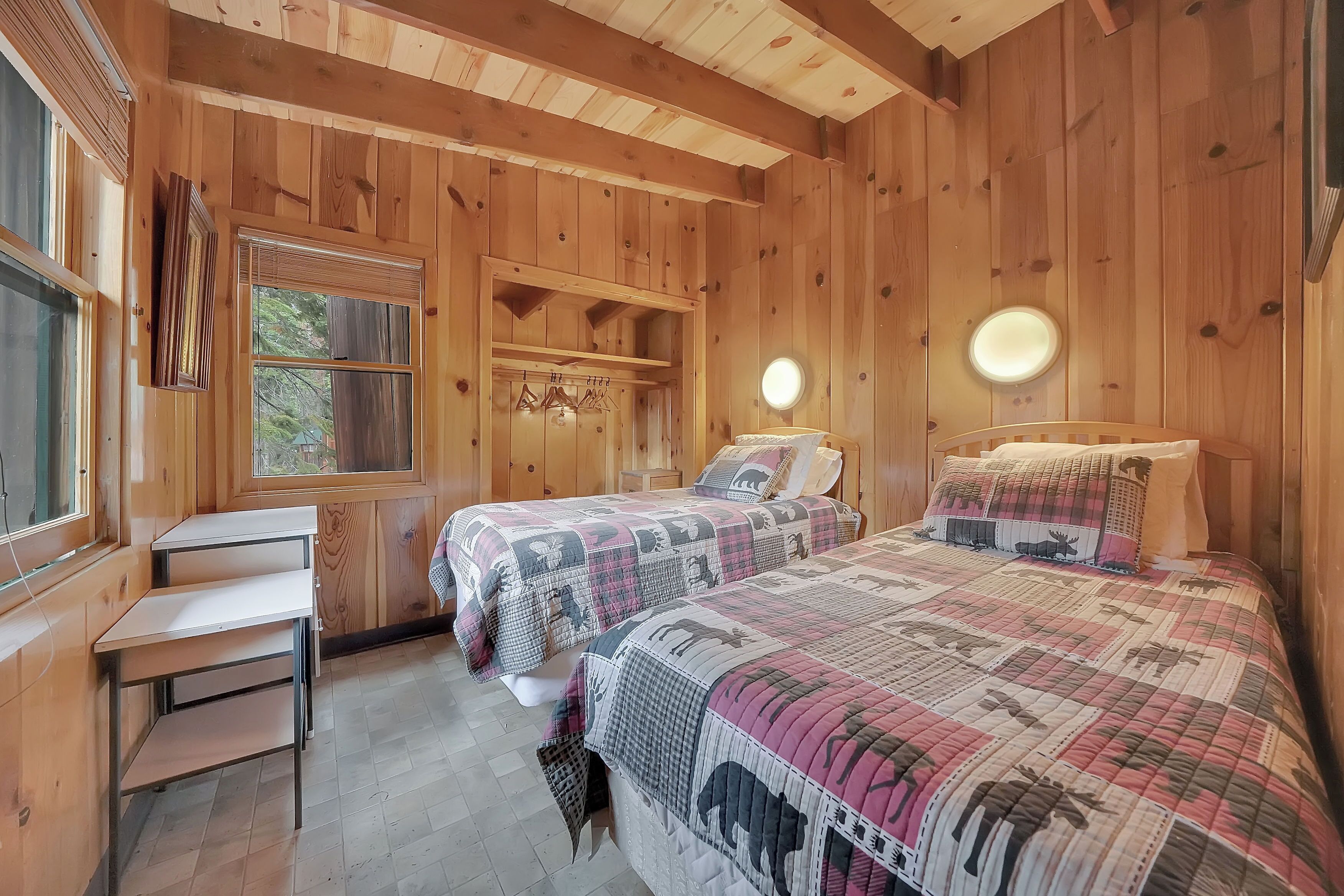 A wooden cabin bedroom with two beds, a desk, and a window.