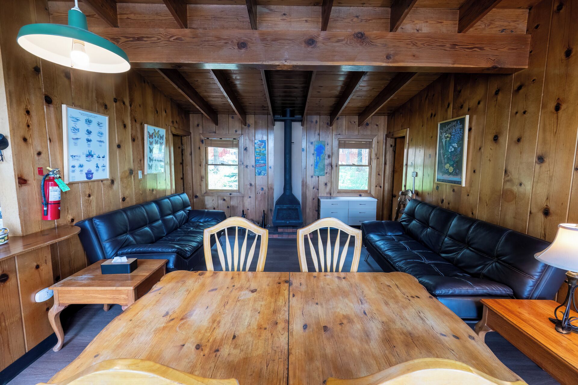 Una habitación acogedora con paredes de madera y una chimenea central. Hay dos sofás de cuero negro frente a una mesa de comedor de madera.