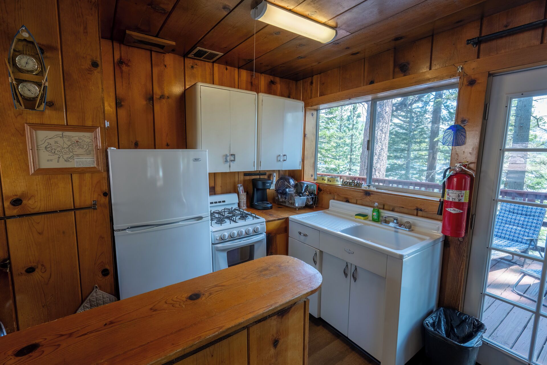A cabin kitchen with wooden walls, a refrigerator, stove, sink, and a window overlooking a forest.