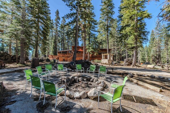 A forested area with a fire pit surrounded by green chairs, a wooden cabin, and tall pine trees. - Kit Carson Lodge (Kit Carson)