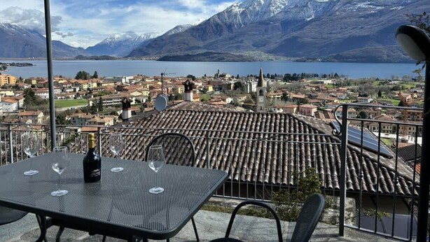 Une terrasse avec une table, des verres à vin et une bouteille, d’où l’on peut admirer une ville entourée de montagnes et bordée d’un lac.