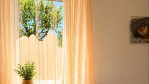 Une pièce avec un canapé, une fenêtre ornée de rideaux légers, une plante en pot et un tableau encadré accroché au mur.