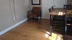 Un salon avec un téléviseur à écran plat fixé au mur, une table à manger en bois avec des chaises, un fauteuil en cuir marron et une porte blanche.