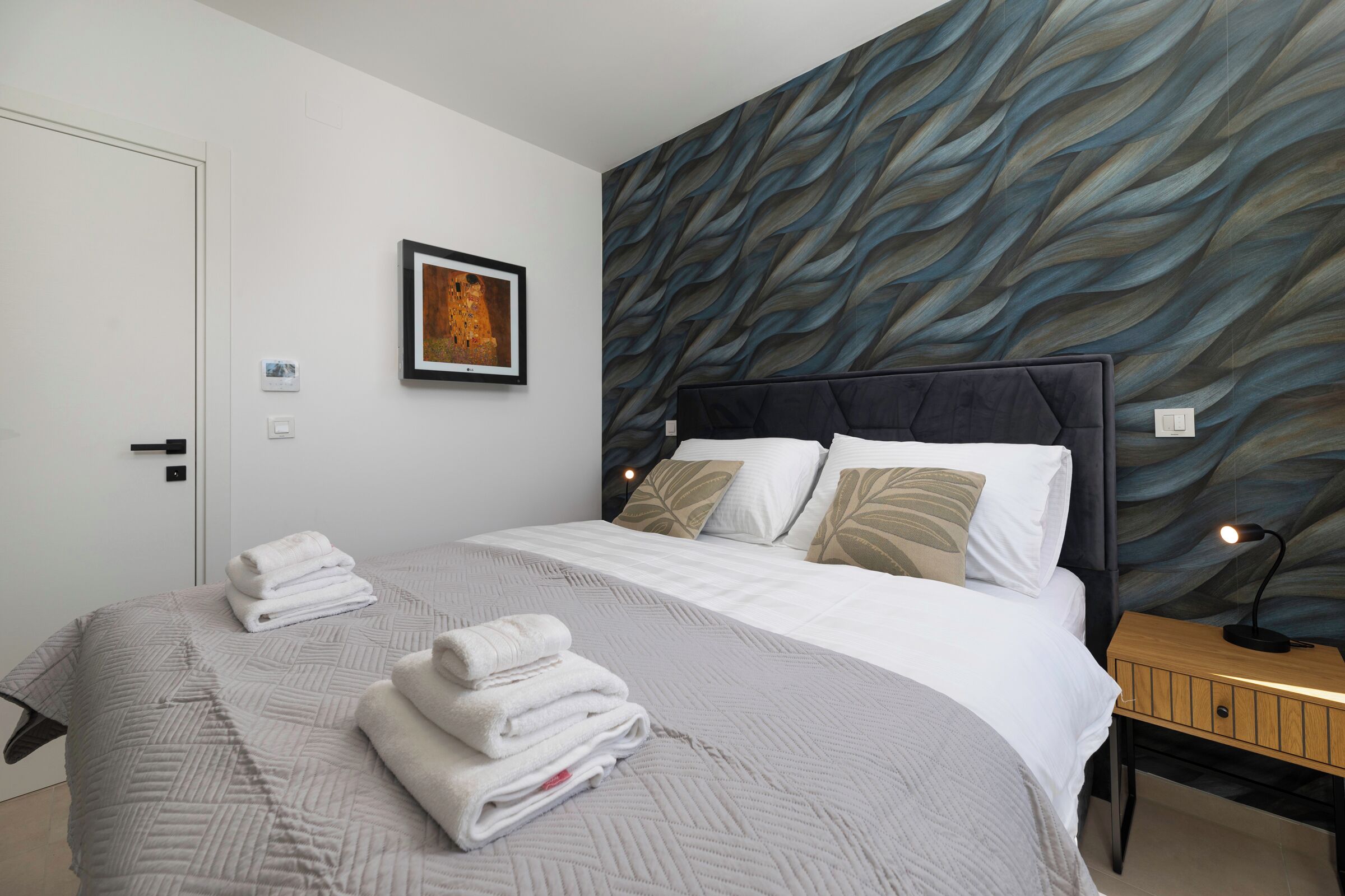 A modern hotel room with a large bed, a headboard with a leaf pattern, a framed artwork on the wall, and a neatly made bed with folded towels.