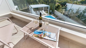 En balkon med gelænder af glas, et bord med et magasin, to vinglas og en vinflaske.