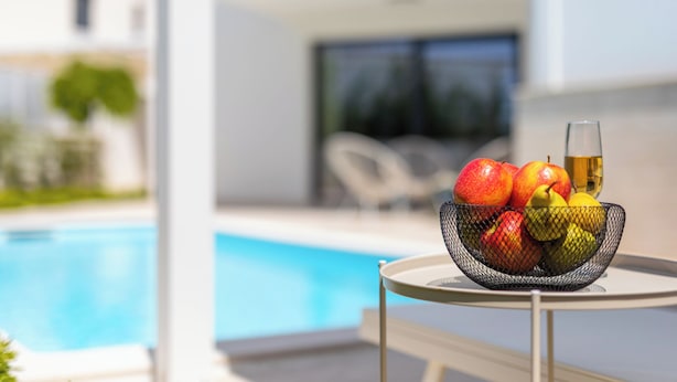 Et rundt bord med en frugtkurv og et glas vin i nærheden af en pool.