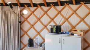 Een kamer met een wit kastje, een waterkoker, een glas en een houten plank met kopjes en een doos.
