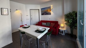 A compact living space with a dining table set for two, a red sofa, a small side table with a lamp, and a large potted plant. - The Wave Musso - Larihome A87 (Musso)