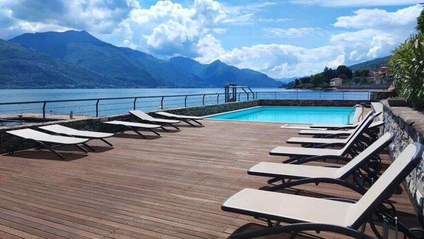 Terraza de madera con tumbonas y piscina, con vista a un lago y montañas al fondo.