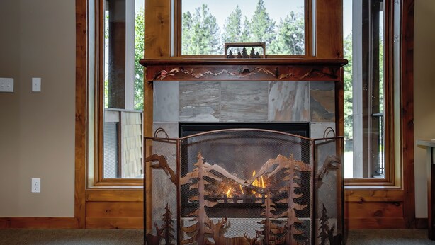 Une cheminée agrémentée d’un paravent décoratif représentant une scène de forêt, située dans une pièce aux murs en bois et aux grandes fenêtres.