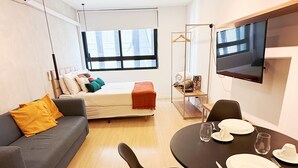 A modern, well-lit room with a bed, a sofa, a dining table set for two, and a TV mounted on the wall.