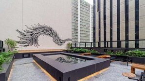 A rooftop terrace with a modern design, featuring a black rectangular pool, seating areas, and a large abstract mural on the wall.