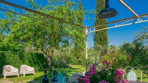 Un área de comedor al aire libre con una mesa preparada para dos, que incluye botellas y copas de vino, rodeada de abundante follaje y un cielo azul despejado.