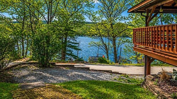 A lakeside property with a wooden deck, a paved pathway, and lush greenery. - Keuka Lakefront | Dock w/ Boat Access | Kayak (Hammondsport)