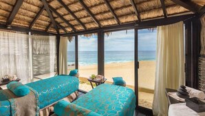 Ein Zimmer am Strand mit einem Reetdach, zwei Betten mit blau gemusterten Bettwäsche, Blick auf den Strand und ein kleiner Tisch mit einer Pflanze.