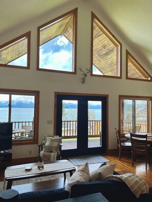 A spacious living room with a large window overlooking a lake, a wooden deck, and a mounted deer head on the wall.
