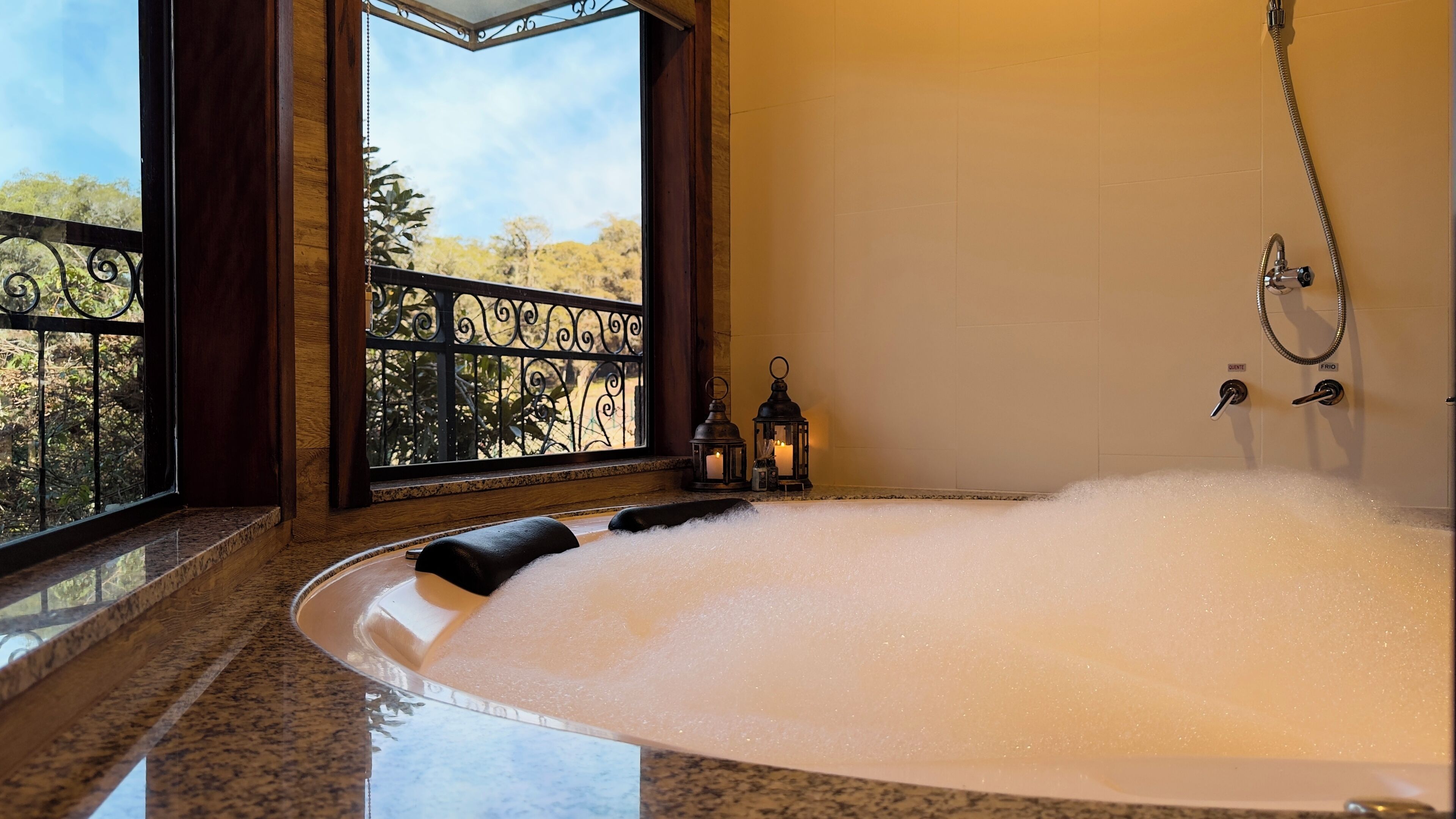 A large, clear bathtub filled with bubbles, positioned next to a window with a view of trees and a metal railing.